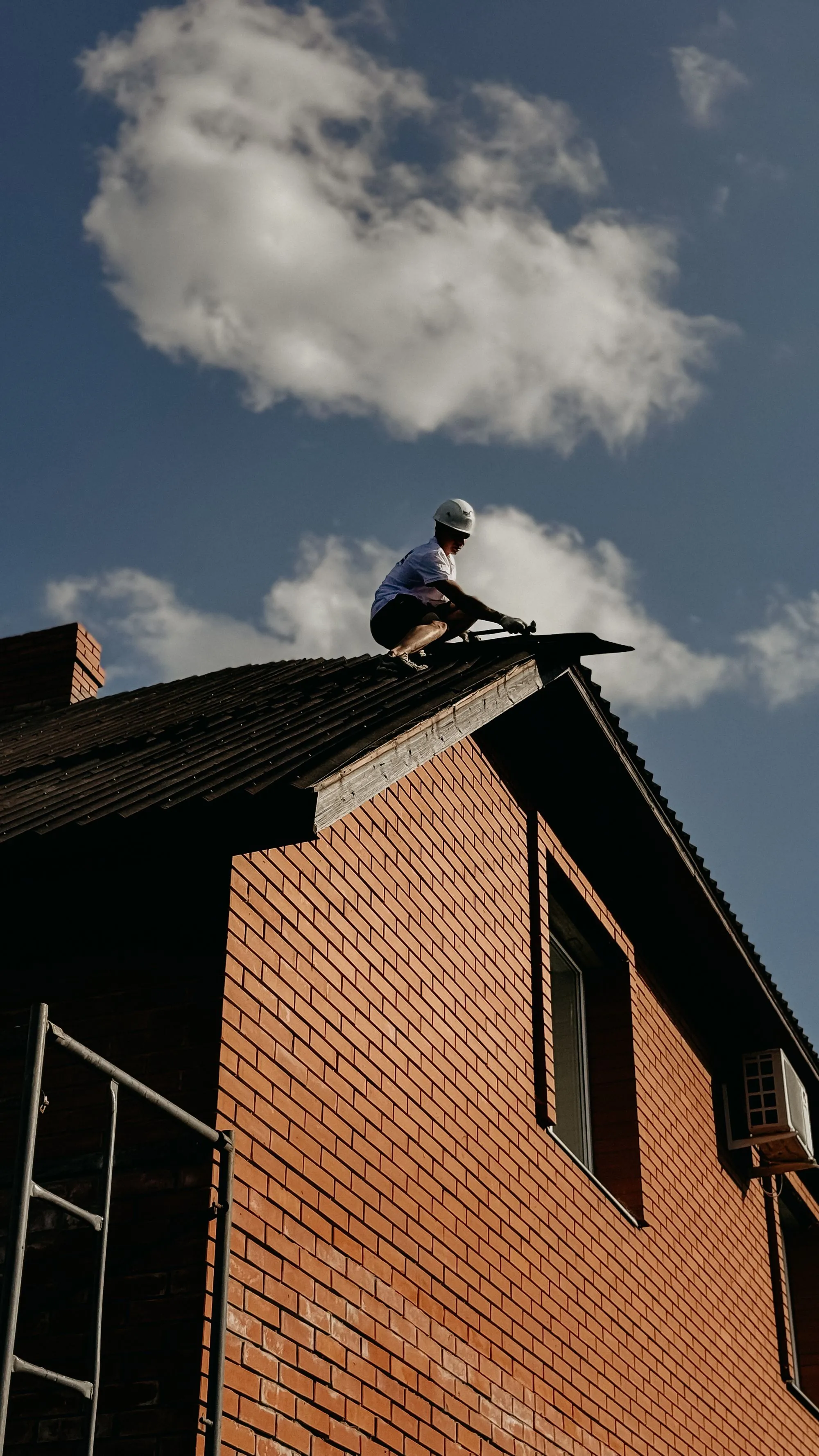 A roofer silhouetted on the ridge of a brick house against the sky