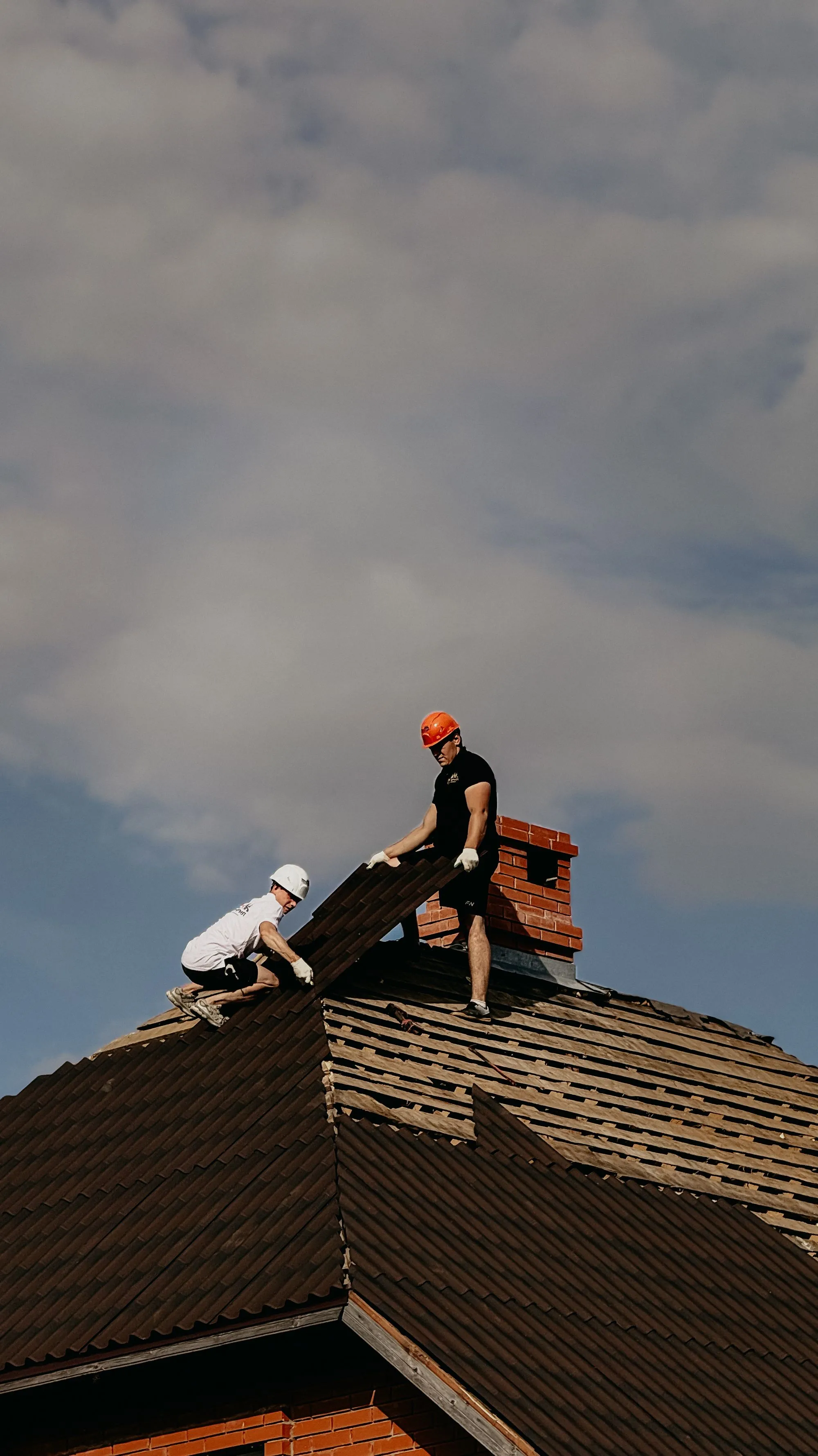 Two roofers lifting a length of material onto a roof beside a chimney
