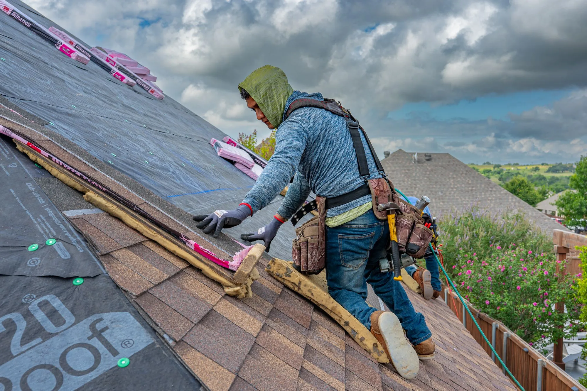 A roofer working a bundle of shingles across a slope under a cloudy sky