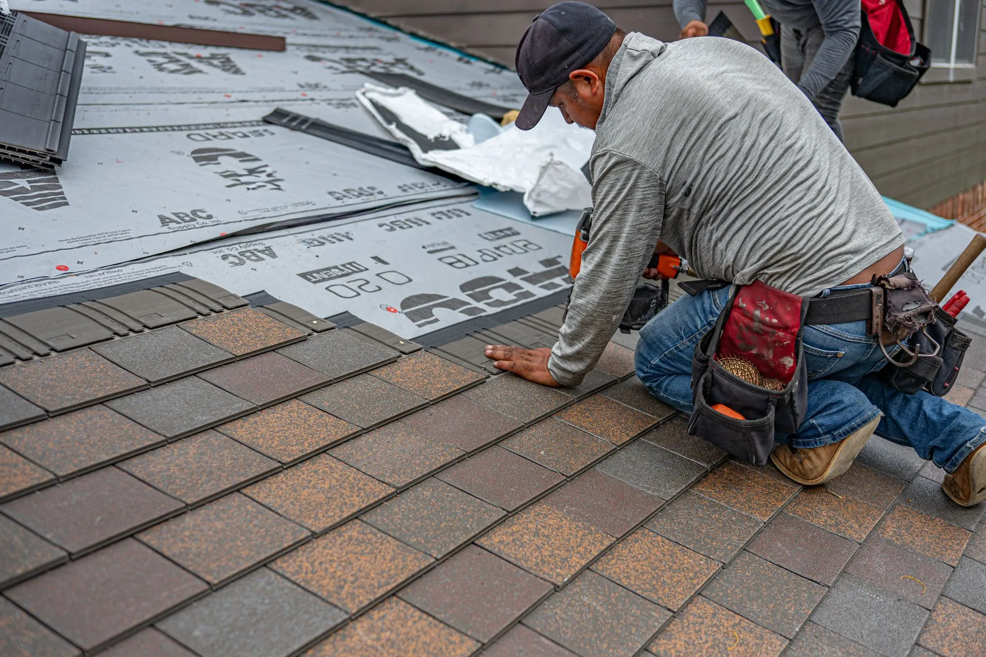 A roofer kneeling to set shingles across a prepared deck