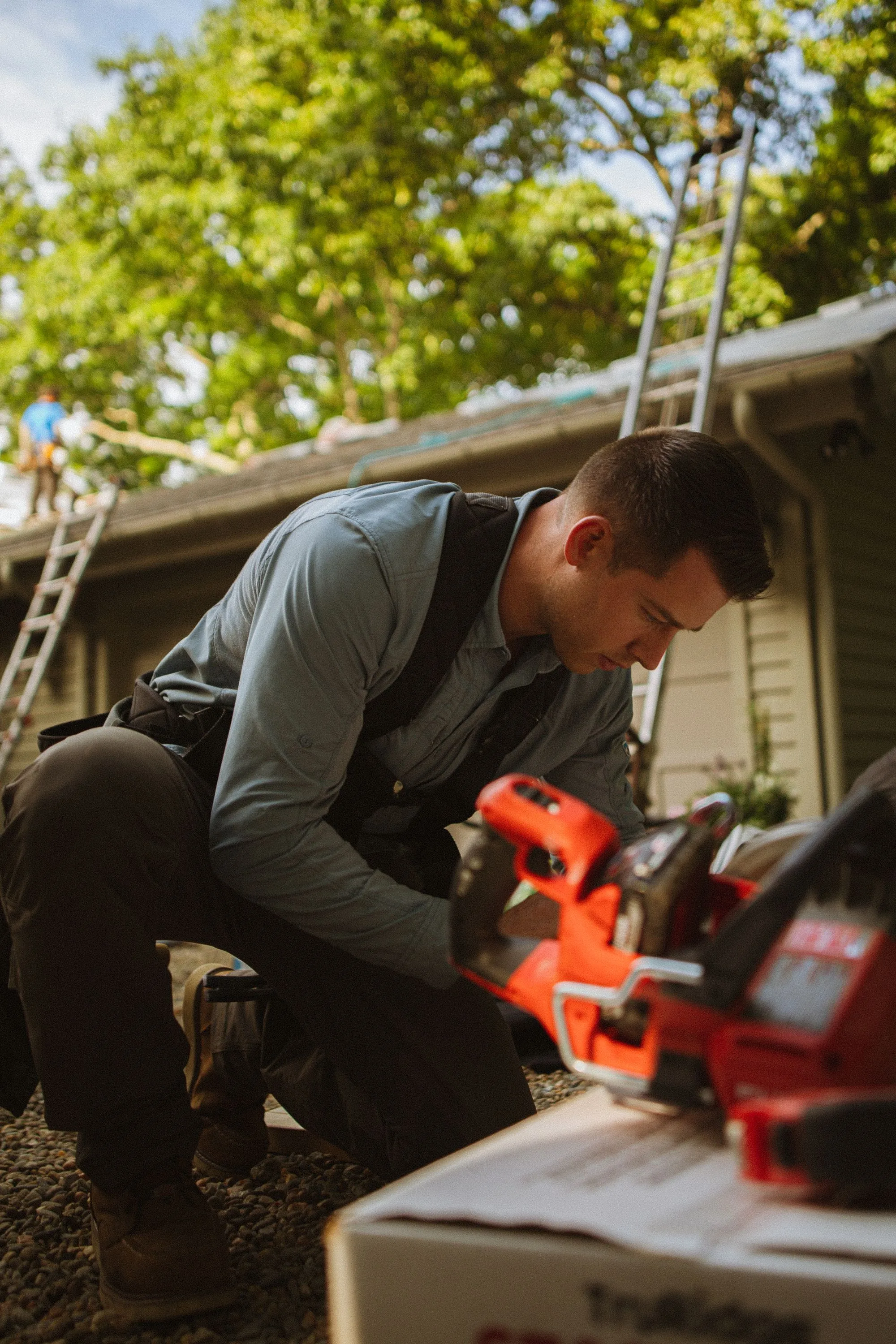 A roofer crouching beside a ladder with his tools at a house