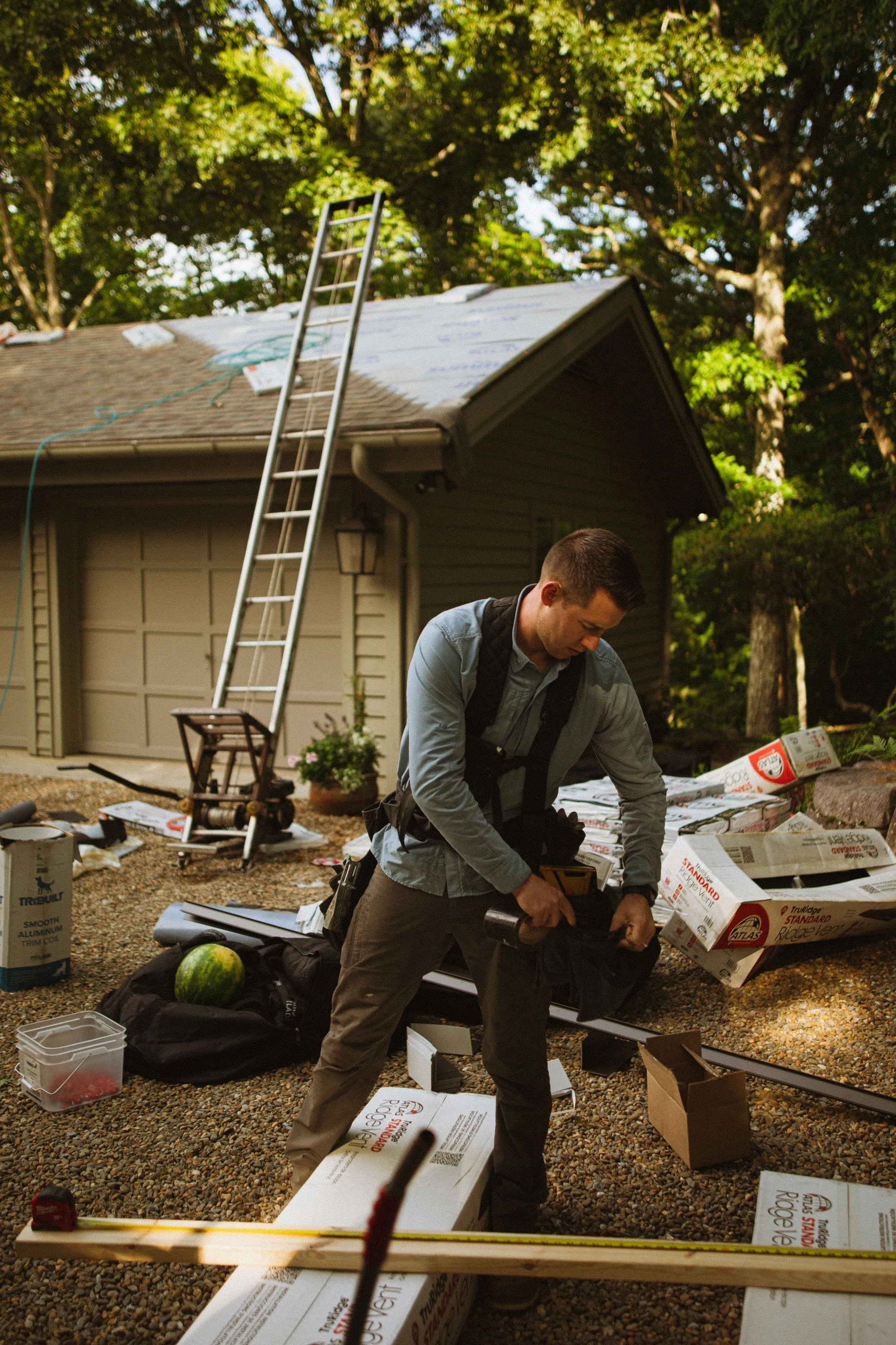 A roofer handling bundles of shingles beside a house with a ladder up